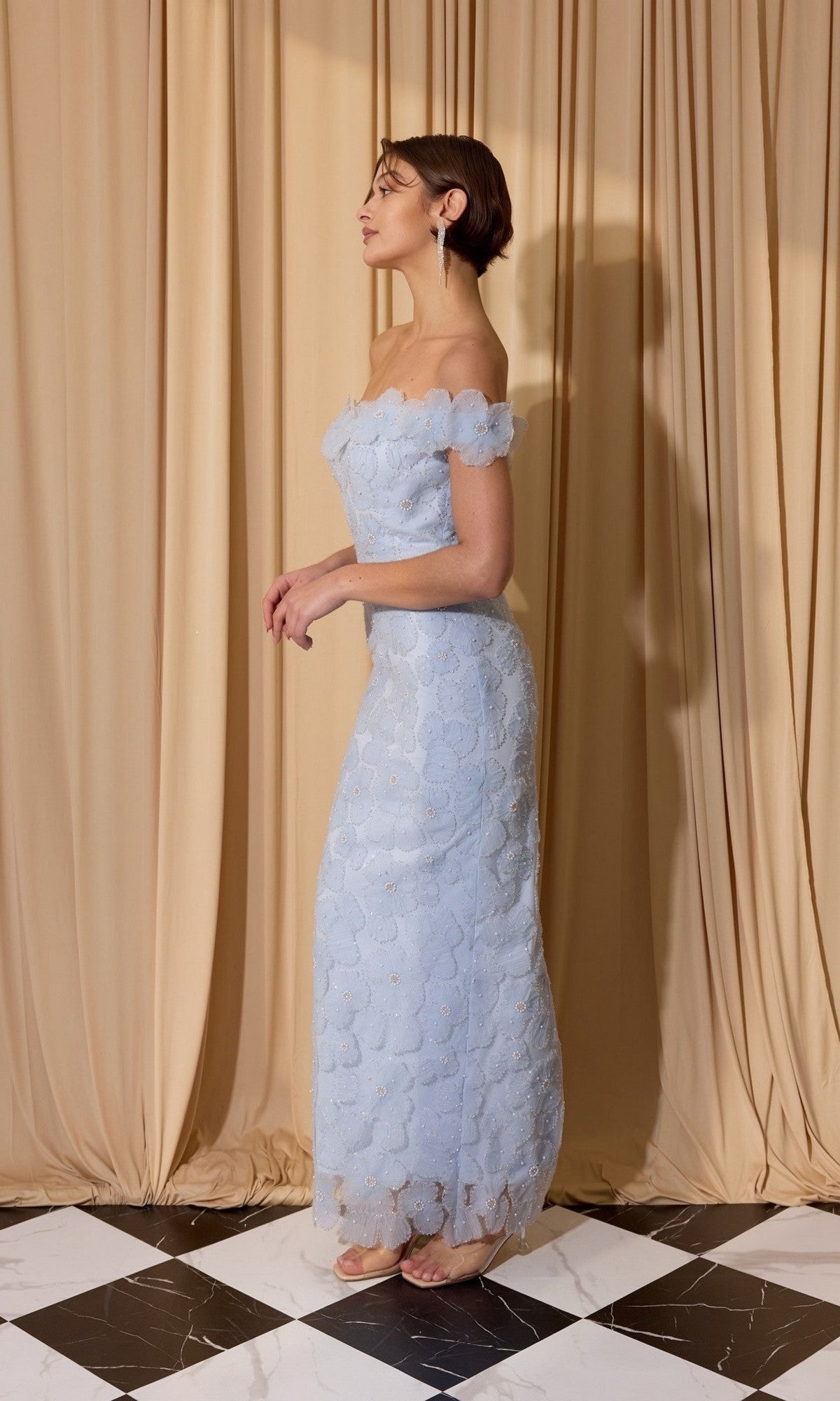 A woman in the Eureka EK205 long prom dress, a light blue off-shoulder lace gown, stands sideways on black and white tiles with beige curtains behind her.